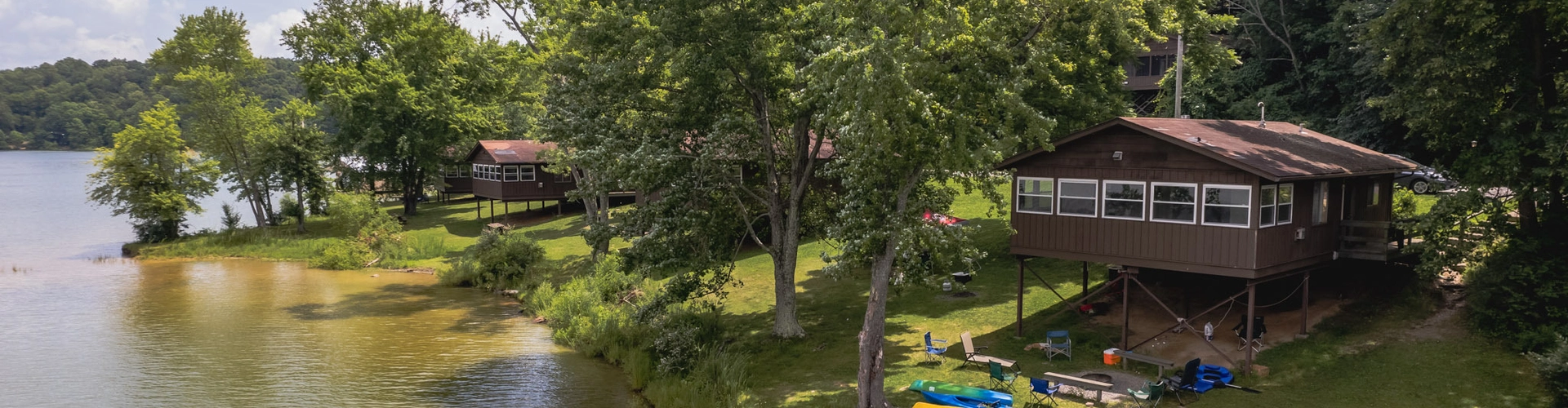 Lakeside cabins surrounded by trees, with kayaks and paddleboards on the grassy shore, and clear water gently meeting the sandy edge under a partly cloudy sky.