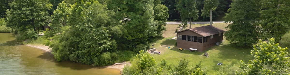 A small brown cabin with a sloped roof sits by the edge of a green, tree-lined lake. The yard is grassy with a fire pit and chairs. Dense forest surrounds the property, and a road runs behind the cabin.