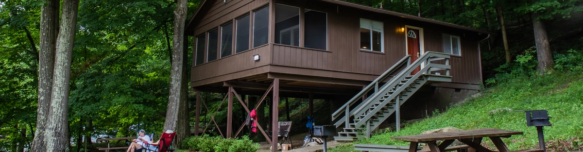 A brown cabin on stilts sits among tall green trees. A picnic table, grill, and fire pit are in front, with a hammock and people relaxing in chairs nearby. Steps lead up to the cabin’s door. The area is lush and shaded.