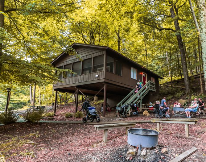 A group of people sits and stands near a brown cabin elevated on stilts in a forest, surrounded by trees. A fire pit and benches are in the foreground, with strollers and outdoor chairs scattered nearby.