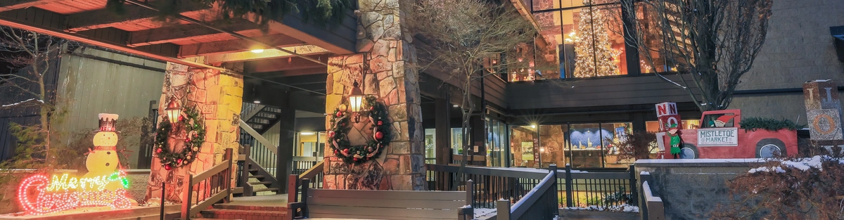 A cozy lodge decorated for Christmas, with wreaths on stone pillars, a lit-up "Merry Christmas" sign with a snowman, holiday lights, and some snow on the ground at dusk.