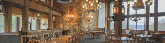 A spacious rustic dining room with wooden tables and chairs, large stone fireplace, chandeliers, high wooden ceiling, and large windows overlooking a scenic outdoor view.