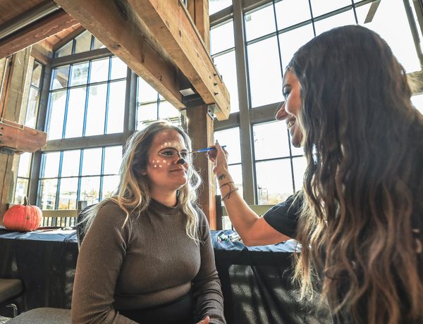 A woman applies face paint to another woman seated inside a rustic lodge with large windows and exposed wooden beams.