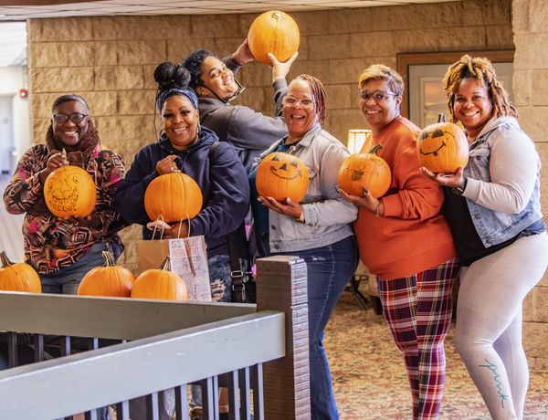 A group of five adults pose with pumpkins at a fall event, with one person holding a pumpkin overhead beside a display of pumpkins in a rustic barn.