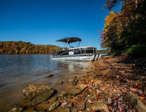 A pontoon boat docked along a rocky shoreline at Salt Fork Lake, surrounded by colorful autumn foliage under a clear blue sky.