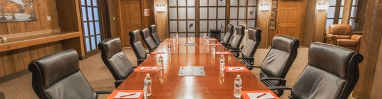 A long wooden conference table with ten black leather chairs, each place set with a red folder, pen, and water bottle. The room features wood-paneled walls, ceiling beams, a chandelier, and framed artwork.