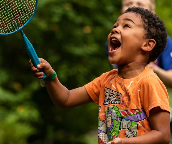 A young child wearing an orange Buzz Lightyear T-shirt excitedly swings a blue badminton racket outdoors, smiling widely with greenery in the background.