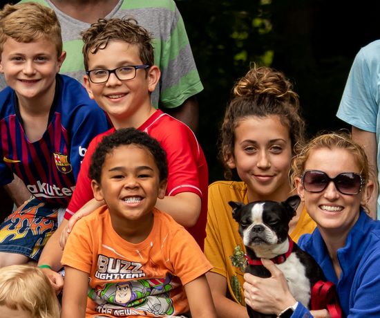 A group of smiling children and a woman wearing sunglasses pose together outdoors; the woman is holding a small black and white dog. Everyone looks happy and relaxed.