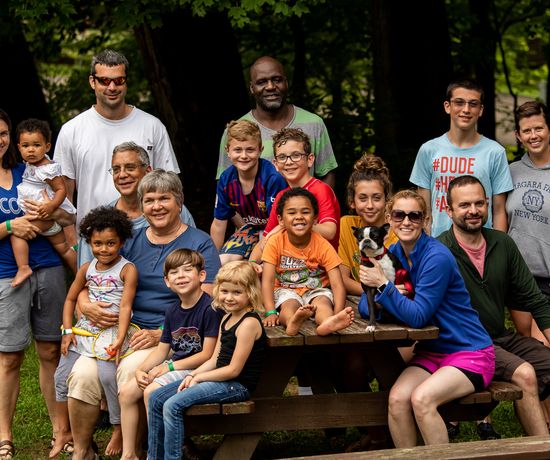 A diverse group of adults and children pose and smile together outdoors around a picnic table, with trees in the background. One person holds a small dog.