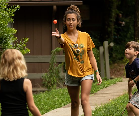 A young woman in a yellow shirt tosses an orange ball to a child with blonde hair while another child looks on, outdoors near a cabin and surrounded by greenery.