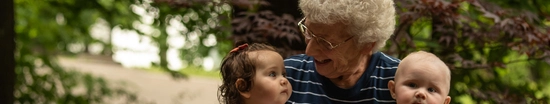 An elderly woman with white curly hair sits on a wooden bench outdoors, smiling as she holds three toddlers on her lap. The background is lush and green with trees, and the group looks relaxed and happy.