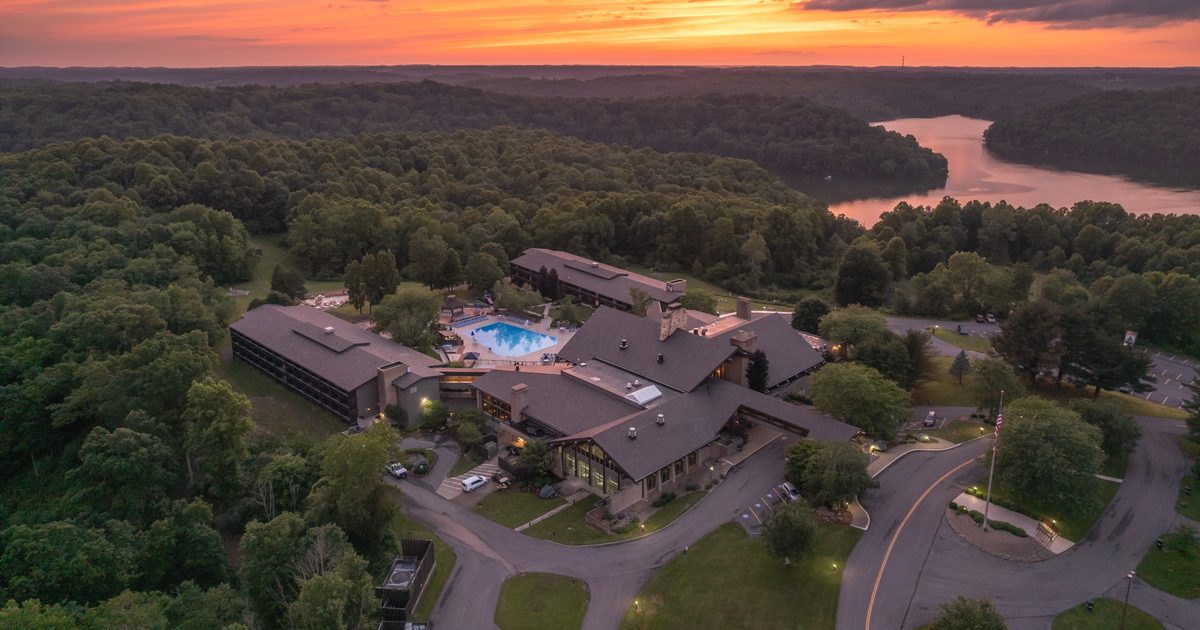 Aerial view of a large lodge with a pool surrounded by dense green forest, roads, and parked cars at sunset, with a river or lake and colorful sky in the background.