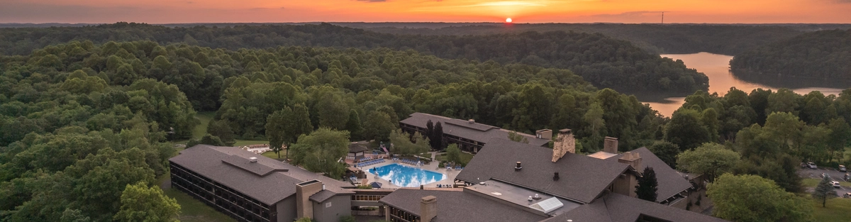 Aerial view of a large lodge surrounded by trees at sunset, featuring an outdoor pool and parking area, with a lake and rolling hills visible in the background.