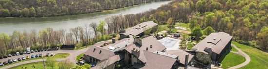 Aerial view of a large lodge-style building surrounded by trees and green lawns near a winding river, with cars parked in multiple lots and forested hills in the background under a cloudy sky.