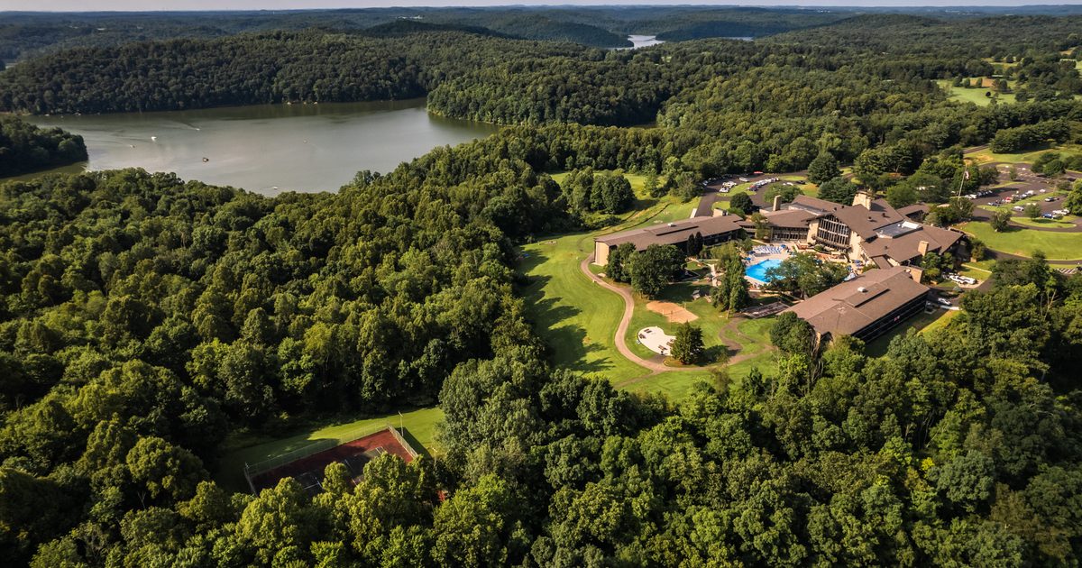 Aerial view of a resort complex with a pool surrounded by dense forest, near a large lake with wooded hills in the background, under a partly cloudy sky.