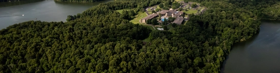 Aerial view of a large building complex surrounded by dense forest and bordered by a winding lake, with green hills and scattered clouds in the background.