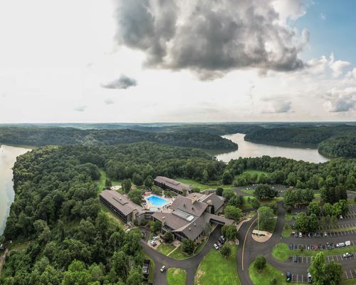 Aerial view of a large resort complex with a swimming pool, surrounded by dense green forests, a winding river, and several parking lots under a partly cloudy sky.