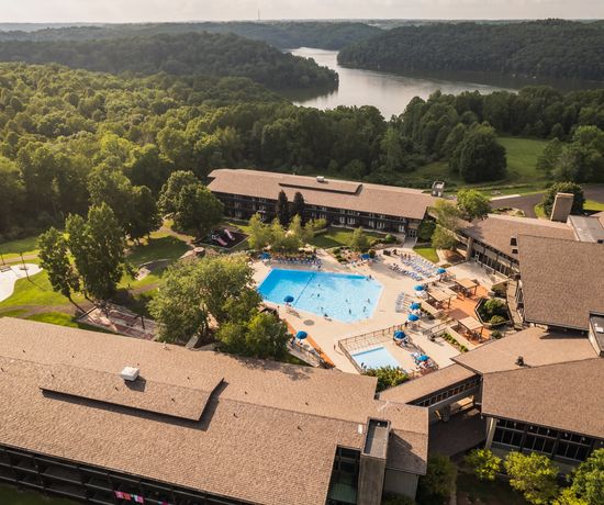 Aerial view of a large lodge surrounded by trees, featuring an outdoor swimming pool, lounge chairs, and a lake in the background with wooded hills under a partly cloudy sky.