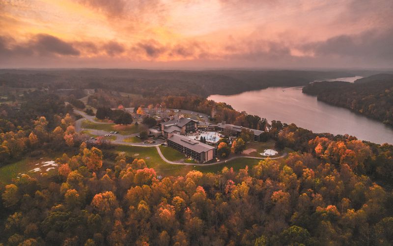 Aerial view of a large building surrounded by autumn trees near a winding river at sunset, with a dramatic sky overhead and rolling hills in the background.