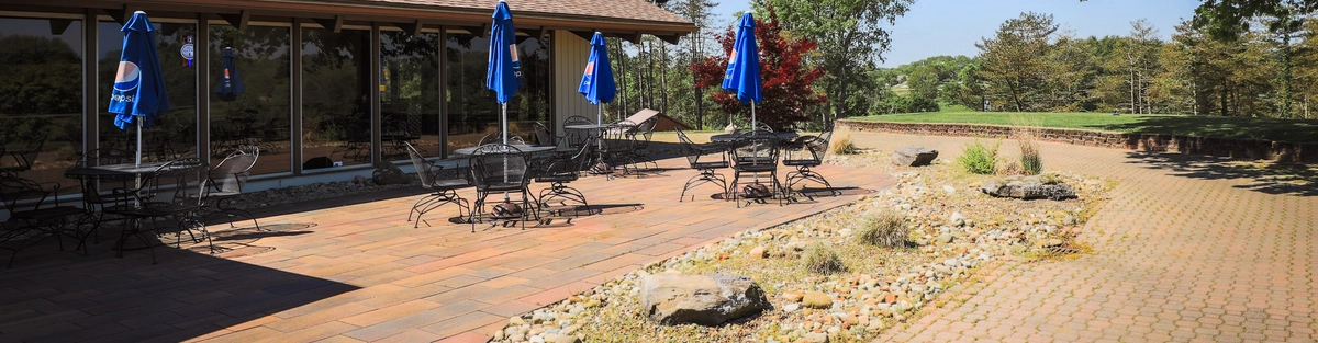 Sunny outdoor patio with empty metal tables and chairs, blue umbrellas, and a brick-paved surface. The area is bordered by trees and shrubs, with a rocky landscaping feature in the center. Building with large windows on the left.