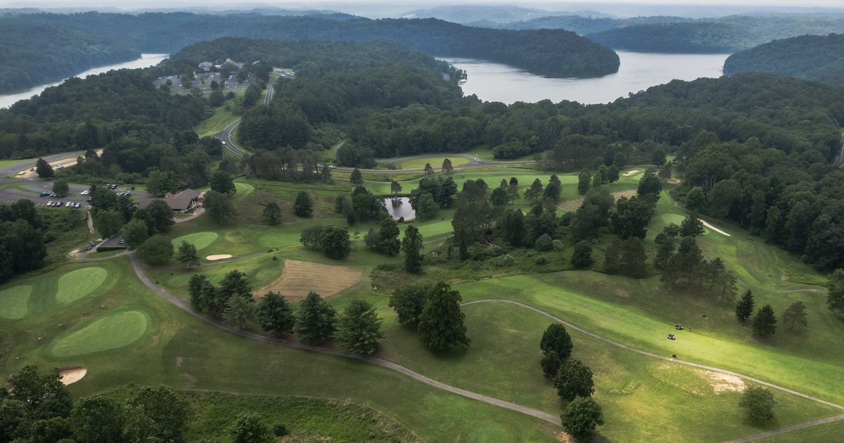 Aerial view of a green golf course surrounded by trees, with a lake and forested hills in the background. A few small buildings and winding paths are visible among the landscape.