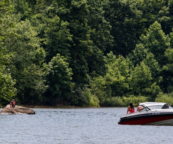 A motorboat with two people floats on a lake surrounded by dense green trees. One person sits on a large rock near the shore, apart from the boat. The scene is calm and summery.