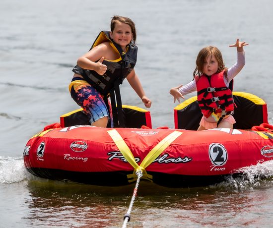 Two children in life jackets ride a red inflatable tube on water, being pulled by a rope. One child is giving a thumbs up and smiling, while the other has their hand raised and mouth open excitedly.