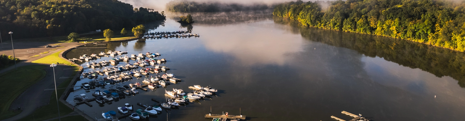 Aerial view of a calm lake with several docks and boats, surrounded by green trees and a grassy shoreline. Morning mist hovers over the water in the distance beneath a clear sky.