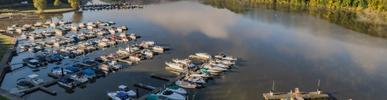 A marina with numerous boats docked in calm water, surrounded by green trees and morning mist, under a clear blue sky. A small dock and building are visible on the right.