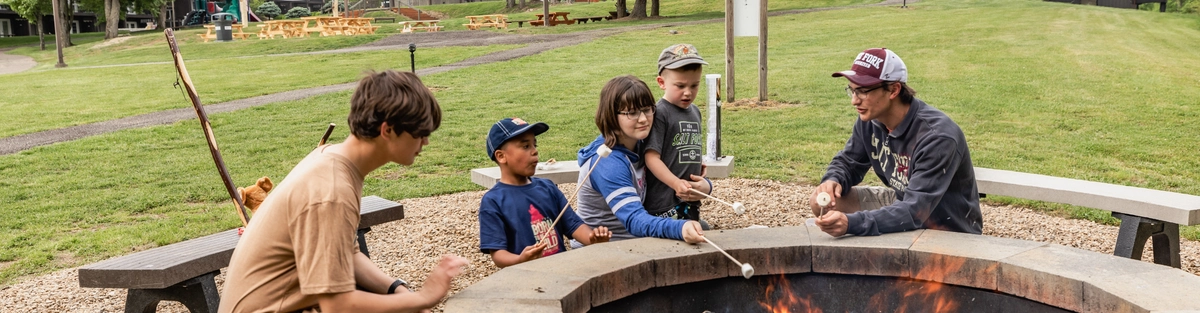 Five children sit around a stone fire pit roasting marshmallows over an open fire in a grassy park, with trees, benches, and buildings visible in the background on a cloudy day.