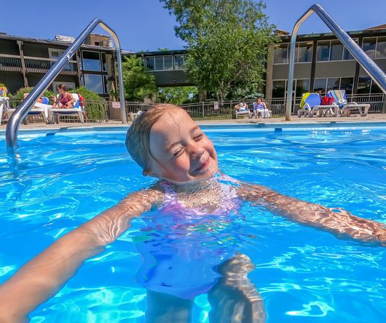 A young child with wet hair smiles while swimming in a bright blue outdoor pool. The sun is shining, and people relax on lounge chairs near a building in the background.