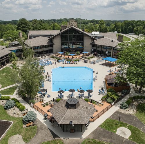 Aerial view of a resort with a large outdoor swimming pool surrounded by lounge chairs, umbrellas, and landscaped gardens, with the main lodge building in the background and lush green trees all around.