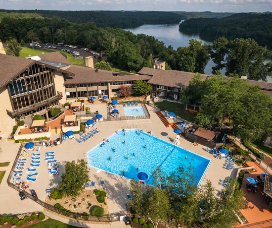Aerial view of a resort with a large outdoor pool, lounge chairs, umbrellas, and surrounding trees; a lake and forested hills are visible in the background under a partly cloudy sky.