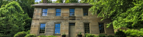A historic two-story stone house with blue-trimmed windows sits surrounded by lush green trees and bushes, under a partly cloudy sky. Stone steps and a path lead to the front door.