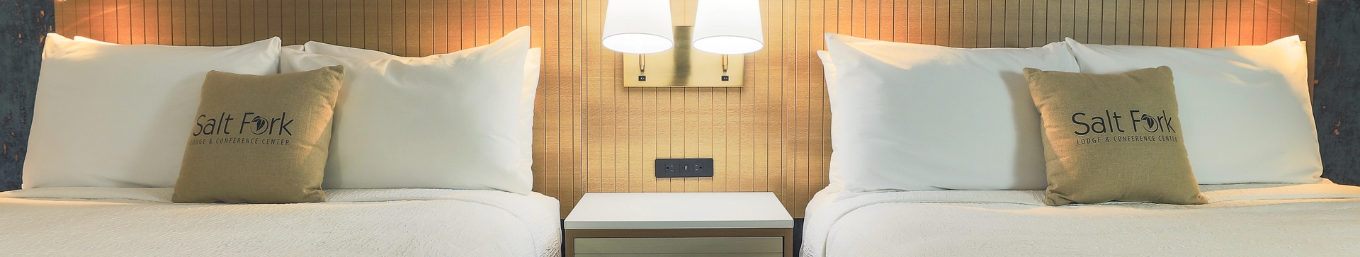 Two neatly made beds with white linens and teal blankets are separated by a small nightstand with two wall-mounted lamps above it. Each bed has a pillow with the text "Salt Fork" on it.