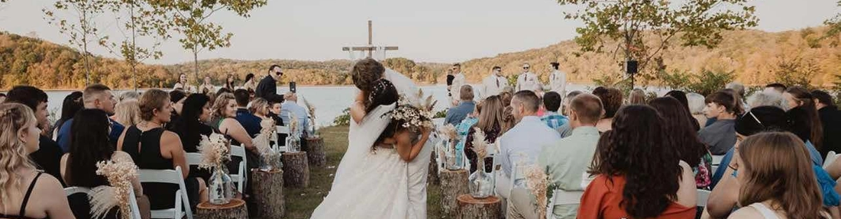Two brides in white wedding dresses kiss at the outdoor altar during their wedding ceremony, surrounded by seated guests. Trees, a large cross, and blue sky appear in the background.