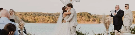 A bride and groom kiss at an outdoor wedding ceremony by a lake, standing before a wooden cross draped with fabric. Guests look on, seated on chairs and tree stumps, with scattered petals on the grass.