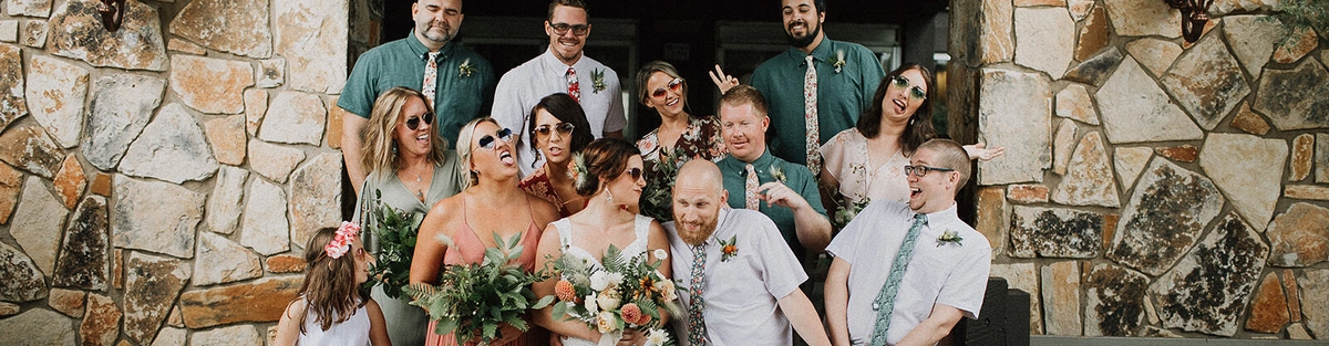 A wedding party poses on stone steps in front of a building. The group includes the bride and groom, bridesmaids, groomsmen, and a flower girl, all smiling and making playful expressions.